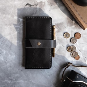 Peut inclure: Un étui de carnet en cuir noir avec un stylo et une fermeture à lanière. Le couvercle a une bordure cousue et un fermoir à bouton en laiton. Un stylo doré est inséré dans la lanière du couvercle. Des pièces de monnaie et un appareil photo sont également visibles.