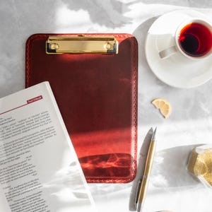Puede incluir: Un portapapeles de cuero burdeos con un clip dorado, junto a un libro abierto, un bolígrafo dorado, una taza de té y una rodaja de limón. El portapapeles es rectangular y tiene un borde cosido.