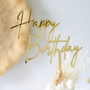 May include: A gold-coloured 'Happy Birthday' cake topper in a cursive font. The topper is placed on a small arrangement of dried flowers and wheat. The background is a blurred, neutral colour.