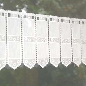Könnte beinhalten: Weiße Spitzen-Fenstergardine mit einem gewellten unteren Rand. Die Gardine hat ein sich wiederholendes Muster aus rechteckigen Paneelen mit dekorativen kreisförmigen und gepunkteten Akzenten. Der transparente Stoff lässt Licht durchscheinen.