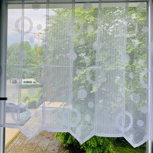 Könnte beinhalten: Weiße, durchsichtige Vorhänge mit einem geometrischen Muster aus Kreisen. Die Vorhänge sind an einem Fenster angebracht und zeigen einen Blick auf Bäume und ein Gebäude in der Ferne.