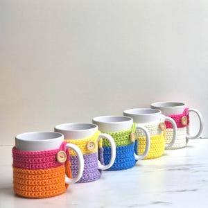 Può includere: Una fila di tazze in ceramica bianca, ognuna adornata con una colorata custodia all'uncinetto. Le custodie presentano una varietà di colori, tra cui rosa, arancione, giallo, blu e verde, e sono fissate con bottoni di legno. Le tazze sono disposte su una superficie bianca.