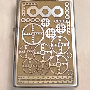May include: A silver-framed lighter with a brass-colored face featuring an engraved steampunk design. The design includes various gears, circles, and rectangular shapes. The lighter is closed and appears to be new.