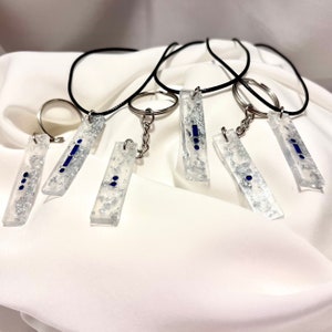 May include: Six silver keychains with clear resin charms. Each charm features a different combination of blue dots and lines. The charms are attached to silver key rings and black cords.