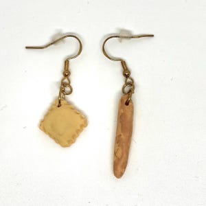 May include: A pair of unique earrings. One earring features a square, light yellow ravioli charm with a scalloped edge. The other earring has a long, brown, breadstick-shaped charm. Both earrings have gold-colored hooks.