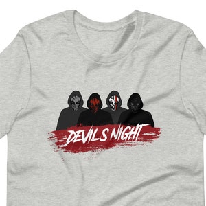 May include: Light gray t-shirt featuring a graphic of four figures in masks above a red brushstroke with the text "DEVILS NIGHT" in white. The figures have varying mask designs, with dark and red accents.