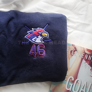 Op de afbeelding: Een marineblauw sweatshirt met een geborduurd logo van een vogel met gekruiste hockeysticks en het nummer 46 in rood en blauw. Een boek met de titel "The Goal" is gedeeltelijk zichtbaar.
