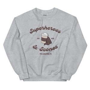 以下が含まれることがあります： グレーのスウェットシャツ。茶色の筆記体で「Superheroes & Scones」と書かれています。コーヒーカップと折りたたまれたナプキンのデザイン。テキストの下には「PHILADELPHIA, PA」と書かれています。