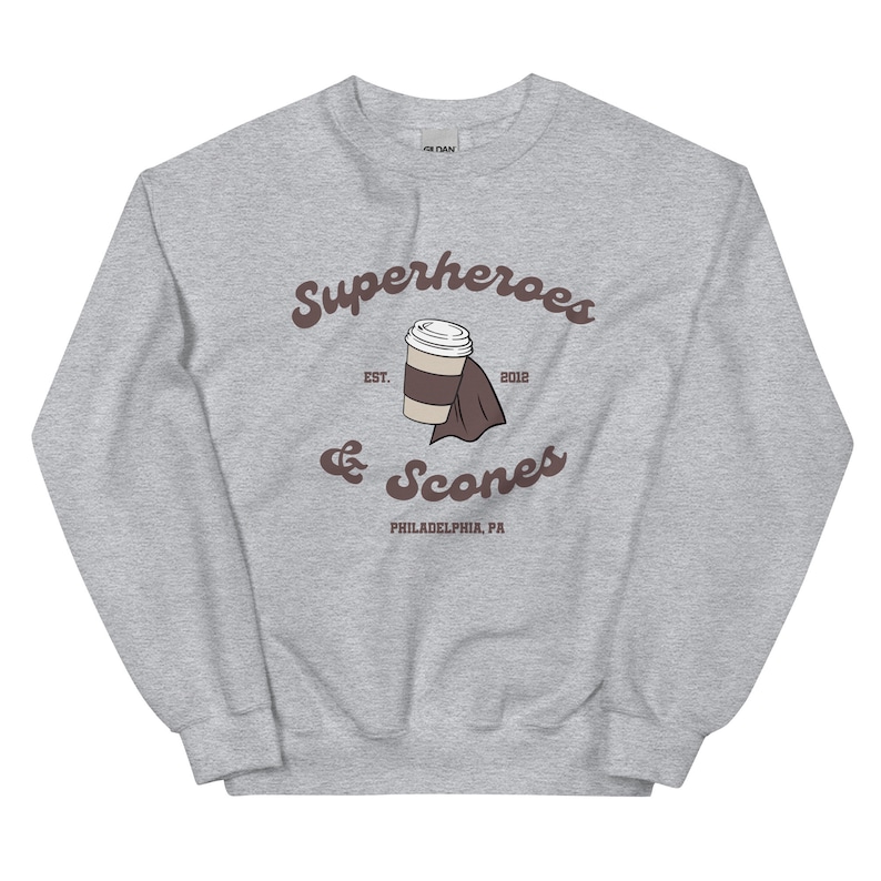 以下が含まれることがあります： グレーのスウェットシャツ。茶色の筆記体で「Superheroes & Scones」と書かれています。コーヒーカップと折りたたまれたナプキンのデザイン。テキストの下には「PHILADELPHIA, PA」と書かれています。