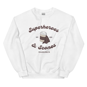 以下が含まれることがあります： 「Superheroes & Scones」の文字とコーヒーカップとスコーンのイラストが描かれた、茶色のグラフィックが特徴の白いスウェットシャツ。「EST. 2012」と「PHILADELPHIA, PA」の文字も含まれています。