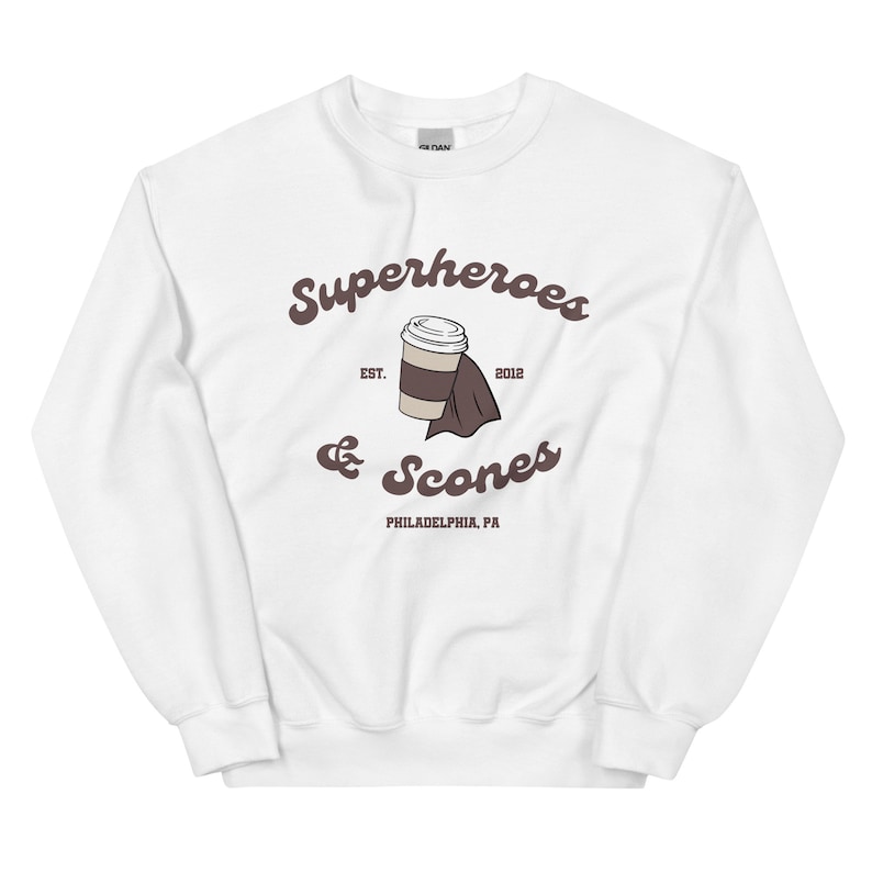 以下が含まれることがあります： 「Superheroes & Scones」の文字とコーヒーカップとスコーンのイラストが描かれた、茶色のグラフィックが特徴の白いスウェットシャツ。「EST. 2012」と「PHILADELPHIA, PA」の文字も含まれています。