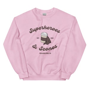 以下が含まれることがあります： 薄いピンク色のスウェットシャツ。茶色の筆記体で「Superheroes & Scones」と書かれています。デザインにはコーヒーカップとナプキンが含まれています。「EST. 2012」と「PHILADELPHIA, PA」の文字もスウェットシャツにプリントされています。