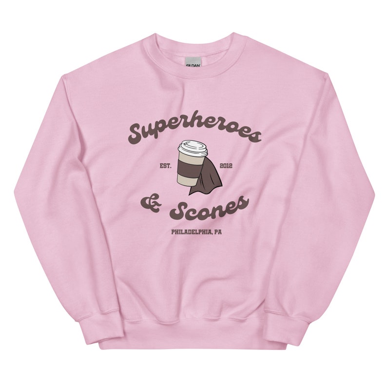 以下が含まれることがあります： 薄いピンク色のスウェットシャツ。茶色の筆記体で「Superheroes & Scones」と書かれています。デザインにはコーヒーカップとナプキンが含まれています。「EST. 2012」と「PHILADELPHIA, PA」の文字もスウェットシャツにプリントされています。