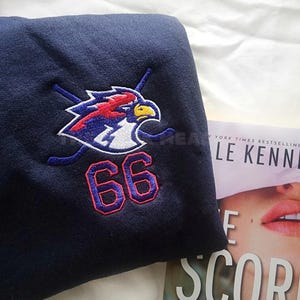 Op de afbeelding: Marineblauw sweatshirt met een geborduurd logo van een vogel en gekruiste hockeysticks, en het nummer "66" in rood en blauw. De sweatshirt is opgevouwen, met een boek gedeeltelijk zichtbaar op de achtergrond.