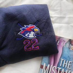 Op de afbeelding: Marineblauw sweatshirt met een geborduurd logo met een gestileerde adelaar, gekruiste hockeysticks en het nummer 22 in rood en blauw. Het sweatshirt is opgevouwen en een boek met de titel "The Mistake" is gedeeltelijk zichtbaar.