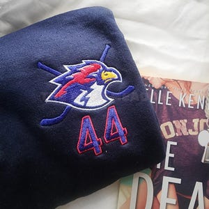 Op de afbeelding: Marineblauwe sweatshirt met een geborduurd logo van een vogel en gekruiste hockeysticks, plus het nummer 44 in rood. De sweatshirt is opgevouwen, met een boek zichtbaar op de achtergrond.