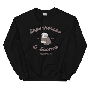 以下が含まれることがあります： スタイリッシュなフォントで「Superheroes & Scones」の文字がプリントされた黒いスウェットシャツ。コーヒーカップとマントのグラフィックも描かれています。「EST. 2012」と「PHILADELPHIA, PA」の文字もプリントされています。