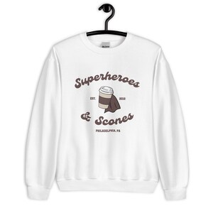 以下が含まれることがあります： 白のスウェットシャツ。茶色の筆記体で「Superheroes & Scones」の文字。中央にはマントをつけたコーヒーカップのグラフィック。テキスト「EST. 2012」と「PHILADELPHIA, PA」も表示されています。