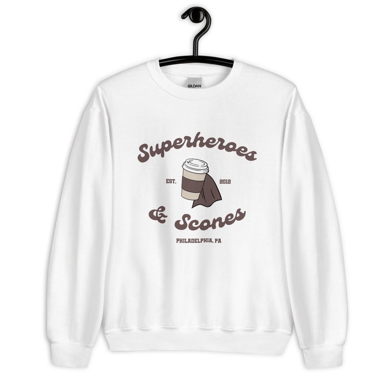 以下が含まれることがあります： 白のスウェットシャツ。茶色の筆記体で「Superheroes & Scones」の文字。中央にはマントをつけたコーヒーカップのグラフィック。テキスト「EST. 2012」と「PHILADELPHIA, PA」も表示されています。