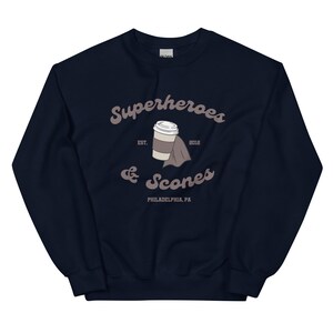 以下が含まれることがあります： スタイリッシュなフォントで「Superheroes & Scones」と書かれたネイビーブルーのスウェットシャツ。デザインには、マントを着たコーヒーカップと「Philadelphia, PA」の文字が含まれています。スウェットシャツはGildanブランドです。