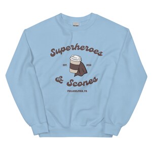 以下が含まれることがあります： 水色のスウェットシャツ。茶色の筆記体で「Superheroes & Scones」と書かれています。デザインにはコーヒーカップとマントが含まれています。「EST. 2012」と「PHILADELPHIA, PA」の文字も表示されています。