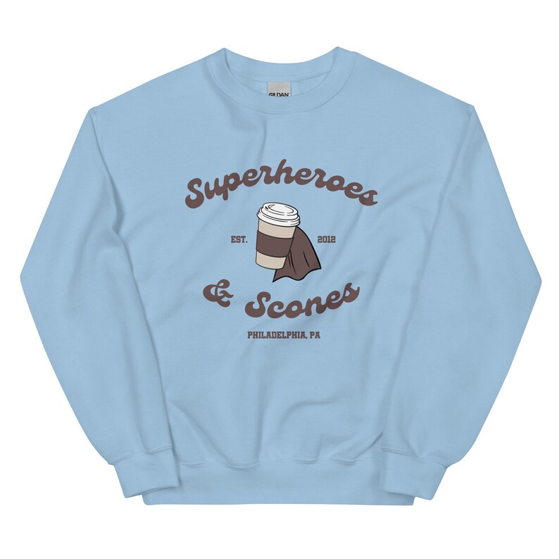 以下が含まれることがあります： 水色のスウェットシャツ。茶色の筆記体で「Superheroes & Scones」と書かれています。デザインにはコーヒーカップとマントが含まれています。「EST. 2012」と「PHILADELPHIA, PA」の文字も表示されています。