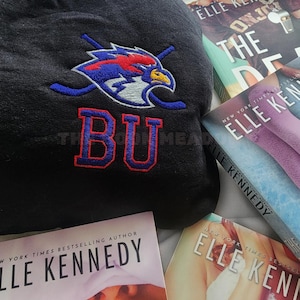 Op de afbeelding: Zwarte hoodie met een rood, wit en blauw geborduurd logo van een vogel en gekruiste hockeysticks, met de letters "BU" in rood. Op de achtergrond zijn boeken van Elle Kennedy te zien.