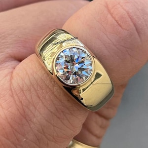 Puede incluir: Un anillo de oro con una gran piedra preciosa redonda y transparente engastada en un engaste de bisel. El anillo tiene una banda gruesa y está hecho de oro de 14 quilates. El anillo está grabado con el texto "CrazyDiamondsCo."