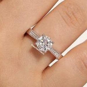 May include: A silver ring with a large, round, clear gemstone as the centerpiece. The band is also adorned with smaller, clear gemstones. The ring is on a person's finger, showcasing its design and sparkle. The ring is a piece of jewelry.