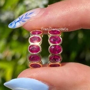 Peut inclure: Deux bagues d'éternité dorées, chacune ornée de pierres précieuses ovales rouge foncé. Les bagues sont présentées sur un fond vert flou. Le texte "CrazyDiamondsCo" est visible.