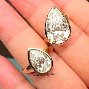 May include: Two gold rings with large, pear-shaped, clear gemstones. The rings are set in gold bands and are displayed on a person's hand. The rings are from CrazyDiamondsCo.