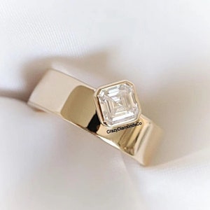 Op de afbeelding: Een gouden ring met een band en een vierkante diamant die in het midden is gezet. De ring ligt op een wit oppervlak. "CrazyDiamondsCo" is op de band gedrukt.