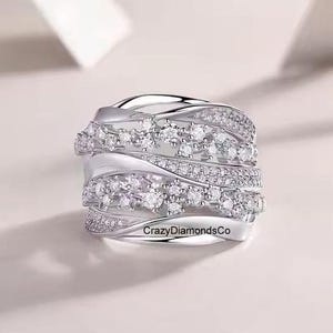Pode incluir: Um anel de prata com várias bandas de pedras de zircônia cúbica brancas brilhantes. O anel apresenta um design ondulado e o texto "CrazyDiamondsCo" está gravado na banda.