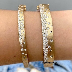May include: Two gold-colored bracelets with a textured finish. Each bracelet is adorned with numerous small, sparkling diamonds, creating a scattered, dotted pattern. The bracelets are worn on a person's wrist.