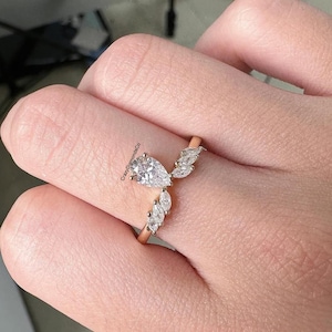 May include: A rose gold ring featuring a pear-shaped diamond center stone, accented by marquise-cut diamonds. The ring's design includes a curved band with the smaller diamonds set along the curve, creating a unique and elegant look.