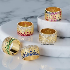 May include: Five gold rings with a textured surface and a band of colorful gemstones. The rings are arranged on a white marble surface. The text "CrazyDiamondsCo" is visible in the image.