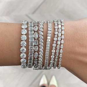 Könnte beinhalten: Eine Auswahl an Diamant-Tennisarmbändern in verschiedenen Designs. Die Armbänder verfügen über runde, smaragd-, marquise- und andere geformte Diamanten. Die Armbänder werden an einem Handgelenk präsentiert und zeigen die verschiedenen Stile und den Glanz. Der Text "CrazyDiamondsCo" ist sichtbar.