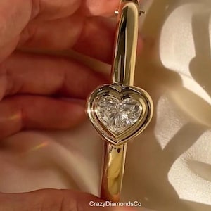 May include: A gold-colored bangle bracelet with a heart-shaped diamond pendant. The diamond is set in a bezel setting within the heart. The bracelet is held against a soft, off-white background. The text "CrazyDiamondsCo" is visible.