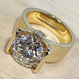 May include: A gold ring with a large, round, clear diamond set in a prong setting. The ring has a thick band and a decorative design on the side. The inscription "CrazyDiamondsCo" is visible on the inside of the band.