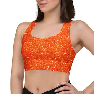 May include: An orange sports bra with a glittery pattern. The bra has a scoop neck and a racerback design. The model is wearing black leggings.