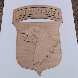 Puede incluir: Una placa de pared de madera de color marrón claro con la insignia de la 101.ª División Aerotransportada. El diseño incluye una forma de escudo con el perfil de una cabeza de águila en el centro y una pestaña arqueada con la palabra AIRBORNE.