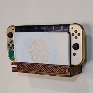 May include: A wooden wall mount for a Nintendo Switch console, featuring a Celtic knot design. The console is gold with a white game card inserted.