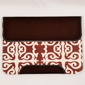 May include: A brown leather laptop sleeve with a white and red patterned design. The sleeve has a flap that closes with a zipper.