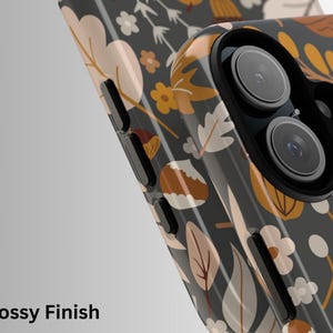 May include: A smartphone case with a glossy finish, featuring a botanical pattern of leaves and flowers in shades of beige, yellow, and white on a dark gray background. The case has a camera lens cutout.