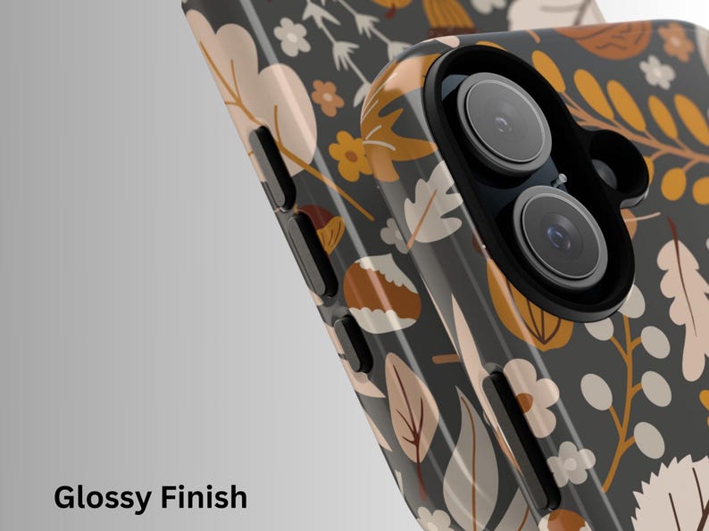 May include: A smartphone case with a glossy finish, featuring a botanical pattern of leaves and flowers in shades of beige, yellow, and white on a dark gray background. The case has a camera lens cutout.