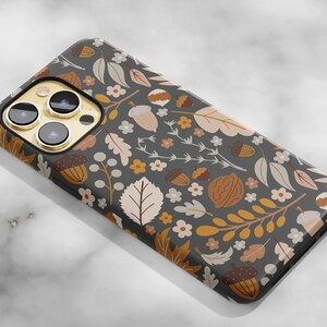 May include: A smartphone case with a dark gray background and a repeating pattern of acorns, leaves, and flowers in shades of brown, tan, and white. The case has a glossy finish and a gold camera lens surround.