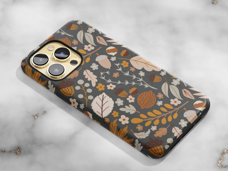 May include: A smartphone case with a dark gray background and a repeating pattern of acorns, leaves, and flowers in shades of brown, tan, and white. The case has a glossy finish and a gold camera lens surround.