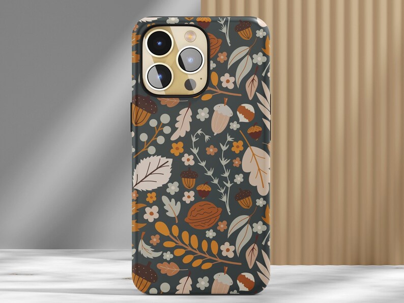May include: A smartphone case with a fall-themed design. The case features a dark gray background adorned with illustrations of acorns, leaves, and flowers in shades of brown, orange, and cream. The case has a glossy finish and a camera cutout.