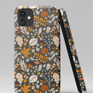 May include: A smartphone case with a fall-themed pattern. The design features leaves, acorns, and walnuts in shades of orange, beige, and white on a dark gray background. The case is shown upright and leaning against a second case.