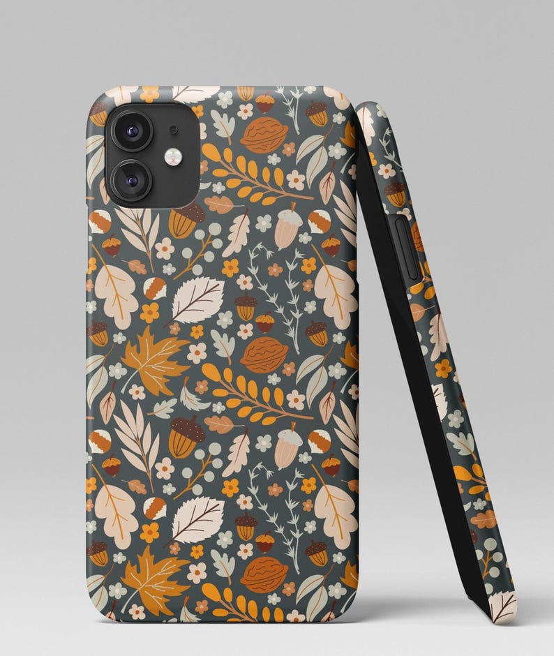 May include: A smartphone case with a fall-themed pattern. The design features leaves, acorns, and walnuts in shades of orange, beige, and white on a dark gray background. The case is shown upright and leaning against a second case.